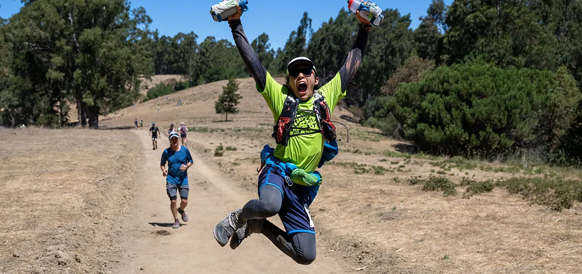 A runner leaps in the air with arms raised in celebration on a dirt trail, with other runners in the background.