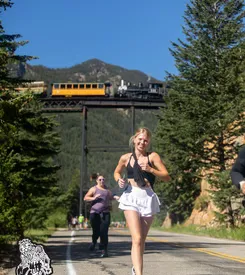 A female runner smiles as she runs on a road with mountains and a train in the background.