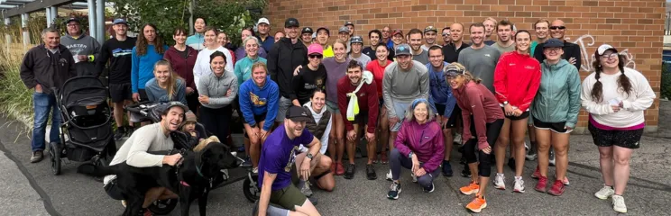 A large group of runners, men and women of various ages, pose for a photo outdoors in front of a brick building. Some are smiling, and one person is holding a dog.