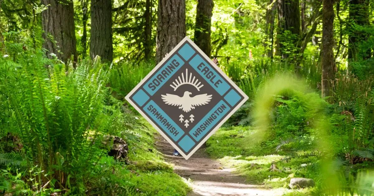 A scenic forest trail with lush ferns and trees, featuring a diamond-shaped race logo for Soaring Eagle Trail Run in Sammamish, Washington.