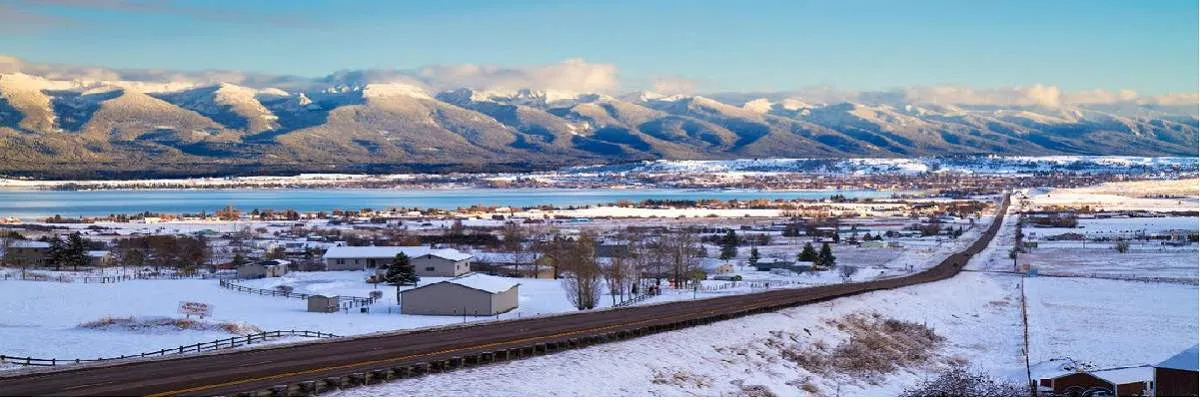 Panoramic view of a snow-covered landscape with a road leading towards a town and a lake, backed by majestic snow-capped mountains under a clear blue sky.