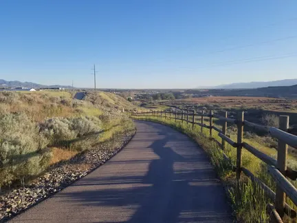 A paved path curves through a landscape with a wooden fence on the right and hills in the distance under a clear blue sky.
