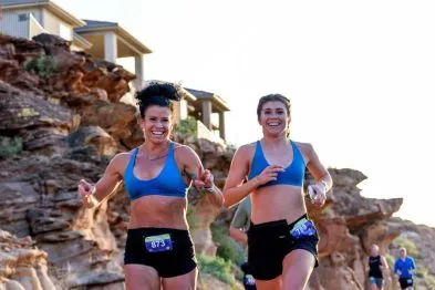 Two female runners smiling as they run on a scenic course with rocky hills and buildings in the background.