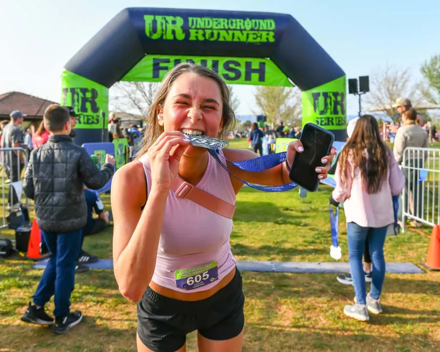 A happy runner celebrates crossing the finish line at the Southern Utah Half Marathon, biting their medal and holding up their phone.