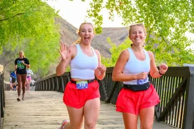 Two female runners in red shorts and white tank tops smile and run across a wooden bridge, with one waving to the camera.