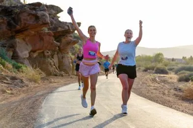 Two female runners with arms raised in celebration cross the finish line of a road race in a scenic desert landscape.