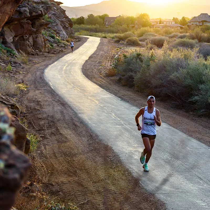 A male runner in a white tank top and black shorts runs on a paved path with desert scenery and mountains in the background during a race.