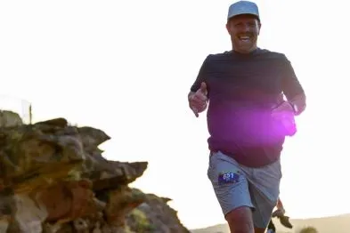 A male runner smiles and gives a thumbs up while running on a rocky path with a bright sky in the background.