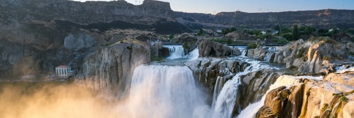 A majestic waterfall cascades down rocky cliffs, with mist rising from the water and a clear blue sky above.