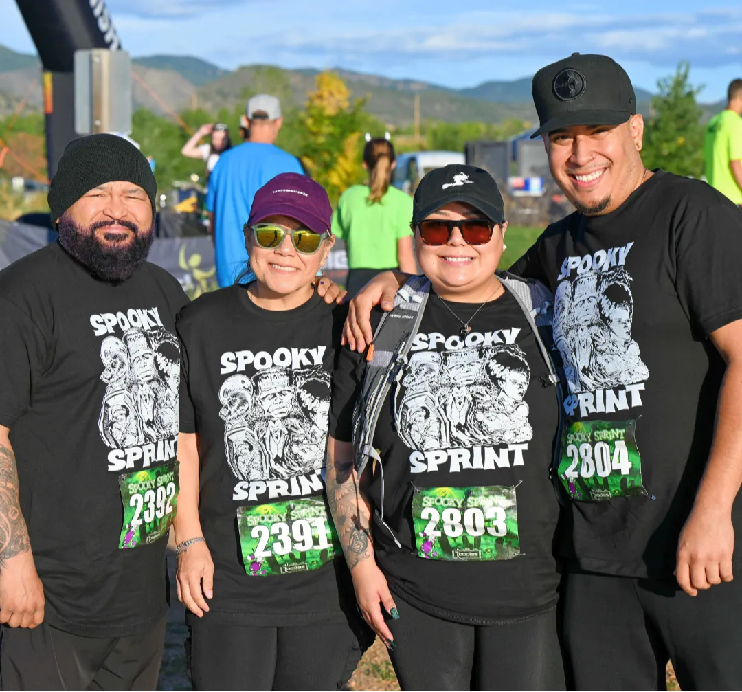 Four smiling participants wearing matching 'Spooky Sprint' t-shirts and bib numbers stand together at the start line of a race.
