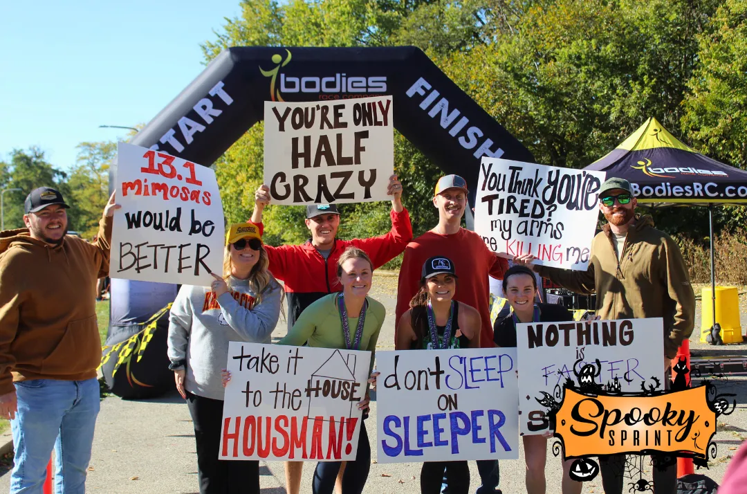 A group of people stand in front of a race arch, holding up humorous signs related to running and the 'Spooky Sprint' race.