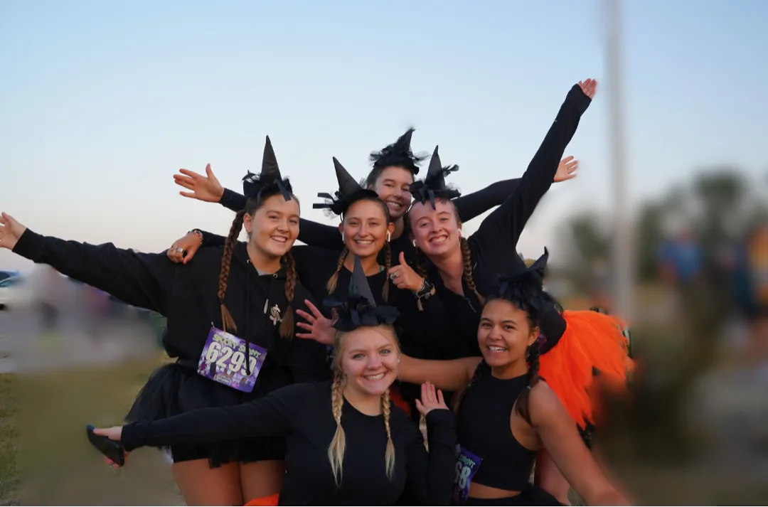 A group of six young women in black witch costumes and hats pose for a photo, smiling and laughing, with race bibs visible.