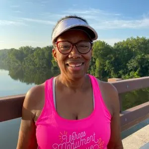 A smiling woman wearing a pink tank top and a visor stands on a bridge with a body of water and trees in the background.