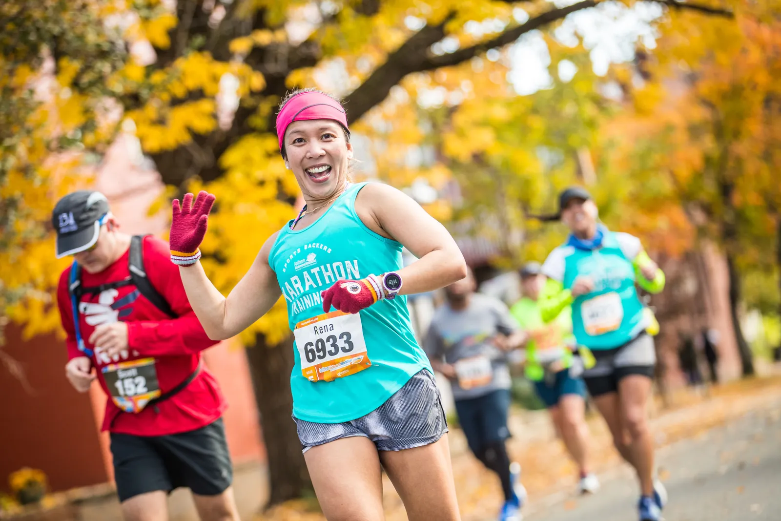 A female runner with a bright smile and a pink headband waves enthusiastically while running in a race, with autumn foliage in the background.