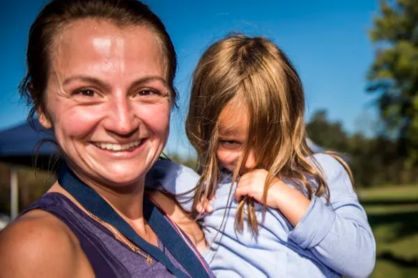 A smiling woman wearing a race medal holds a child who is looking away.