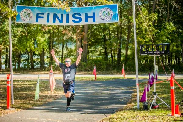 A male runner with arms raised in triumph crosses the finish line at the Sri Chinmoy Half Marathon, with a digital clock showing 1:22:29:51.