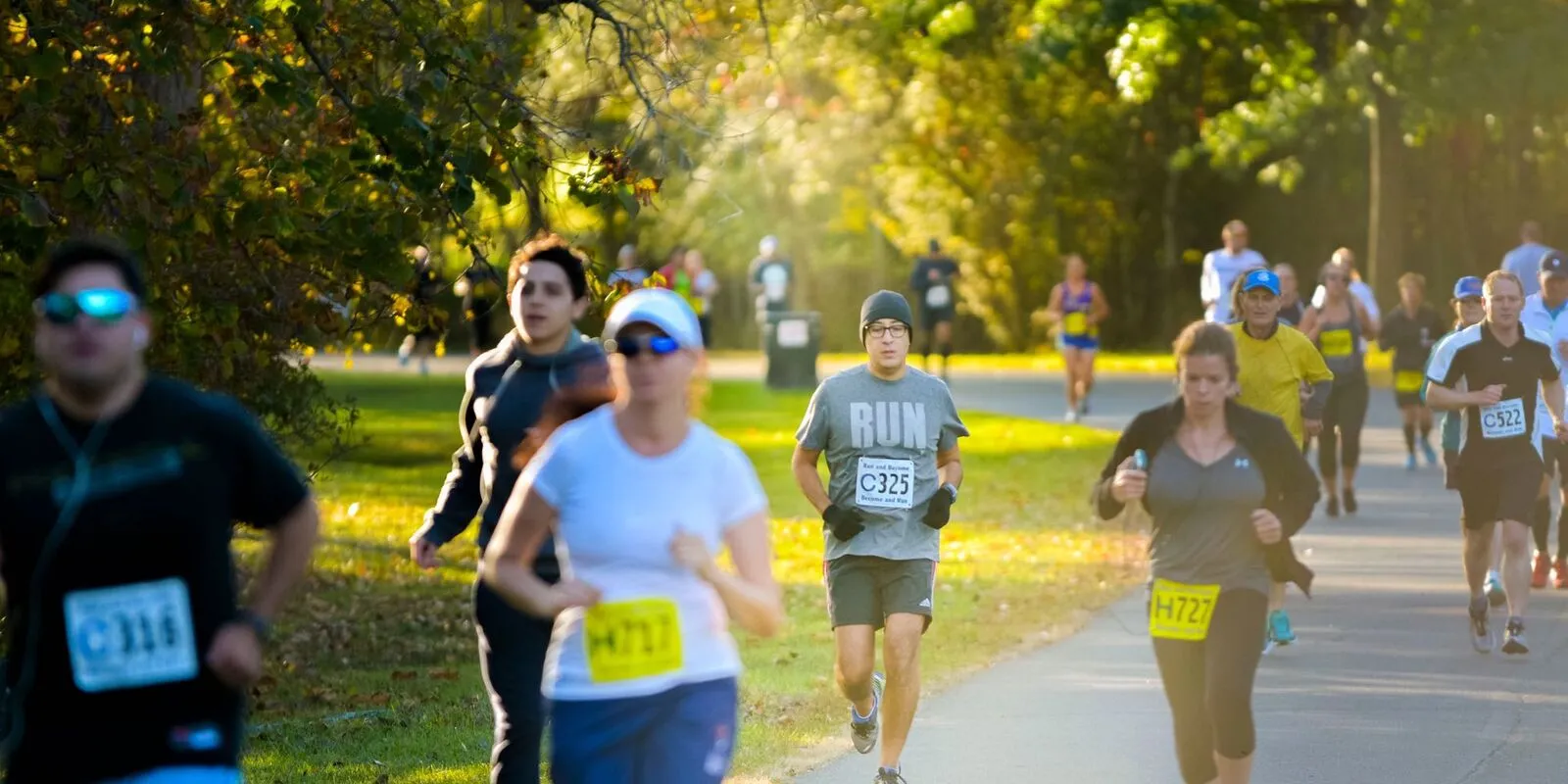 Runners on a paved path in a park, with trees and sunlight in the background.