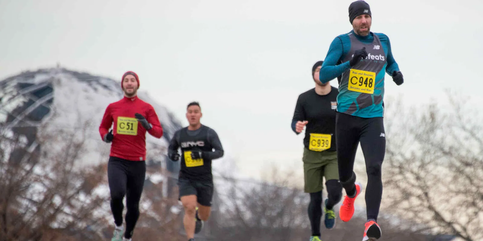 Runners in a race, with bib numbers visible, running on a road with a snowy dome-like structure in the background.