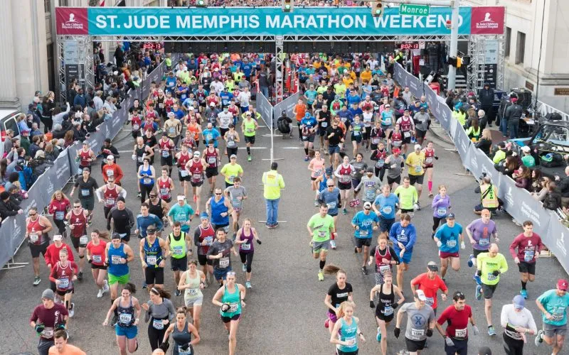 A large group of runners starting the St. Jude Memphis Marathon, with spectators on the side and a banner overhead.