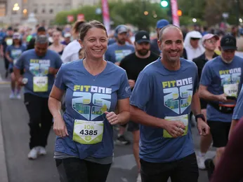 Two runners in matching blue t-shirts with 'FITONE 25' logo smile as they run in a crowd at the start of a race.