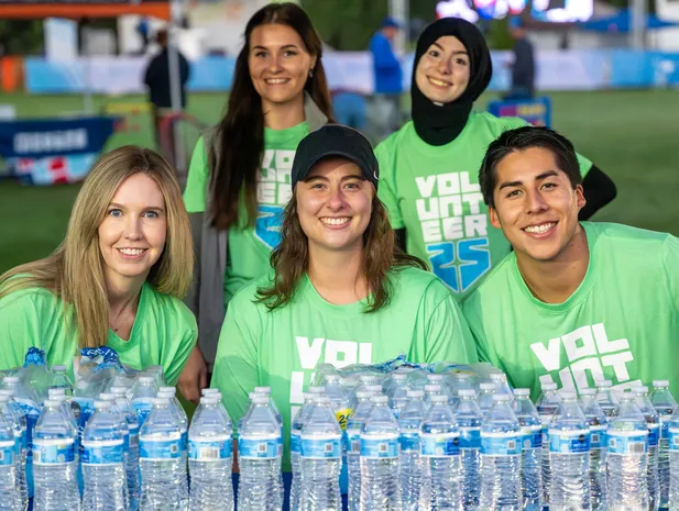 A group of five smiling volunteers wearing bright green t-shirts with 'VOLUNTEER' printed on them, standing behind a table laden with bottles of water.