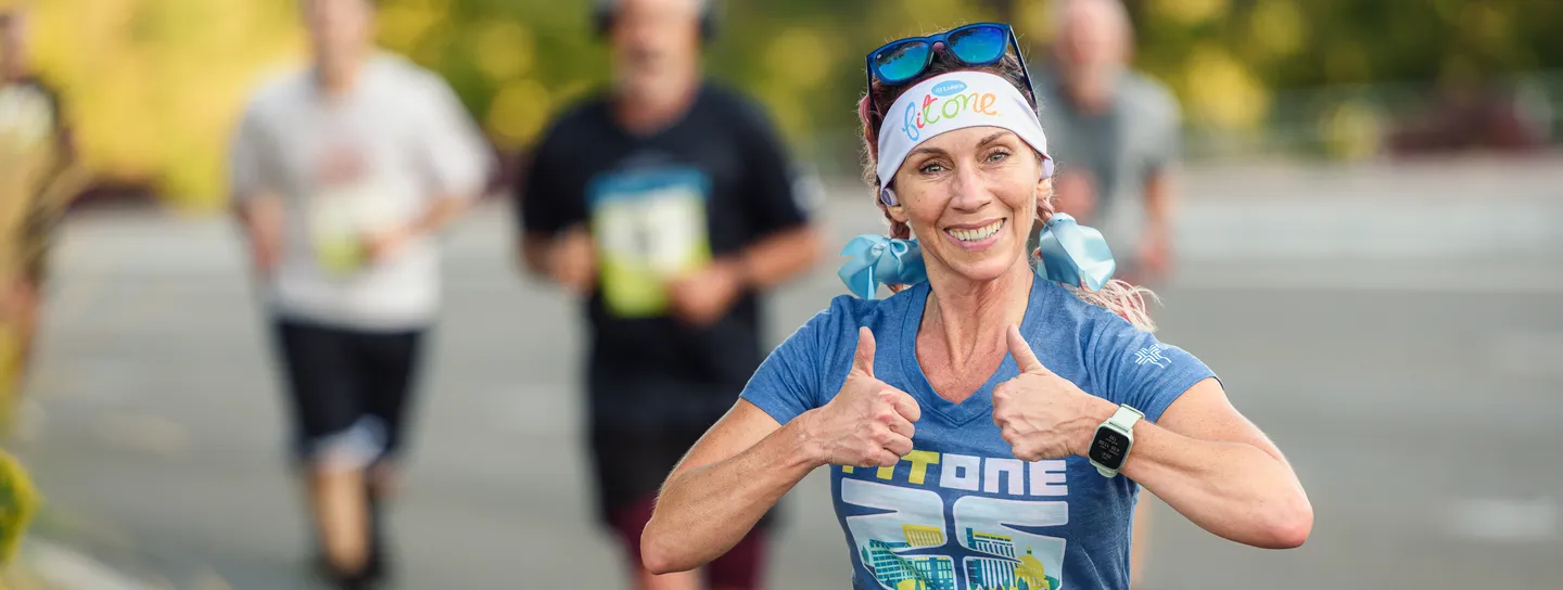 A smiling female runner with pink-streaked hair and blue ribbons in her pigtails gives two thumbs up while running, wearing a blue t-shirt and a watch.
