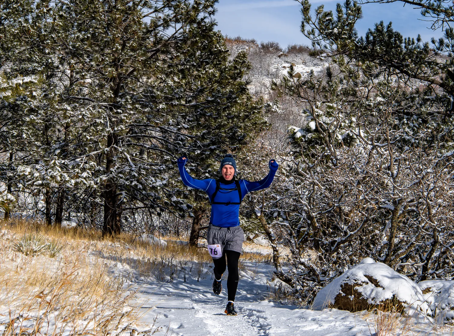 A runner with arms raised in celebration runs on a snowy trail through a forest.