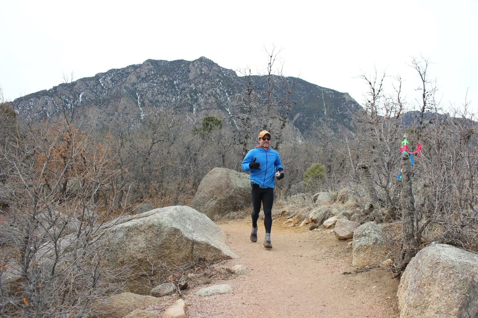 A male runner in a blue jacket and black leggings runs on a dirt trail with mountains in the background.