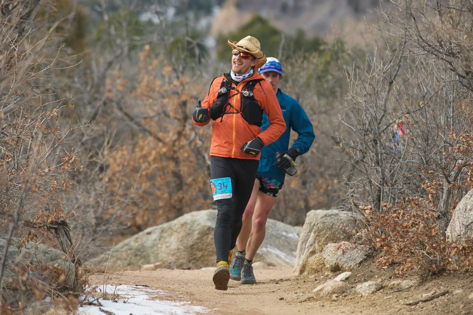 Two runners on a trail, one in an orange jacket and straw hat giving a thumbs up, the other in a blue jacket.