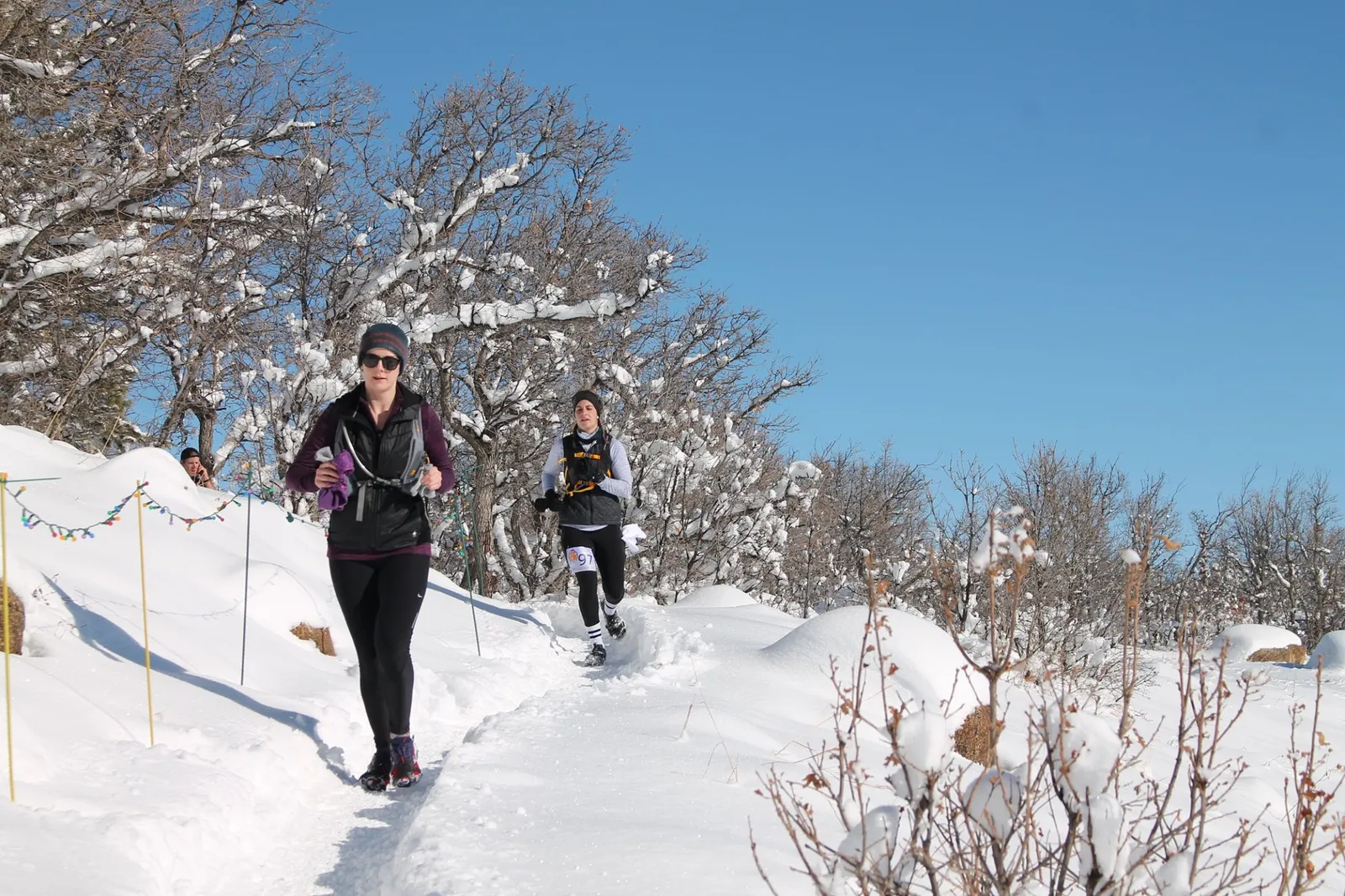 Two runners on a snowy trail with trees in the background.