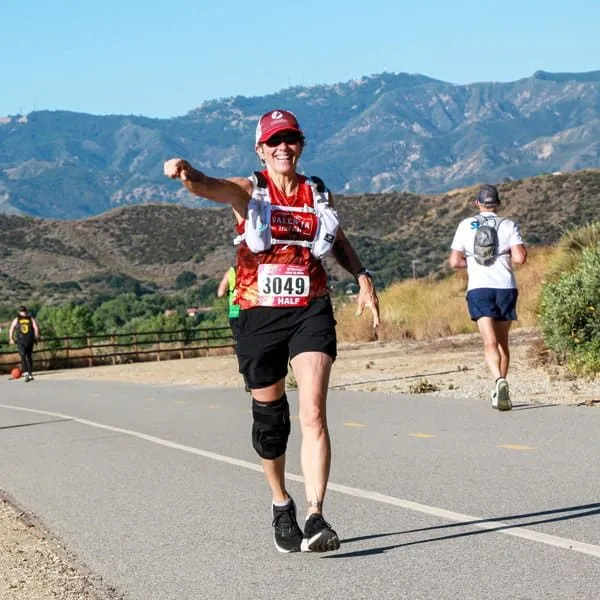 A female runner with bib number 3049 smiles and points forward while running on a paved road with mountains in the background.