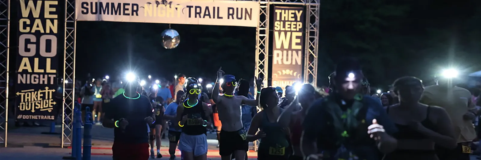 Runners with headlamps and glow sticks starting a night trail race under a banner that says 'Summer Night Trail Run'.
