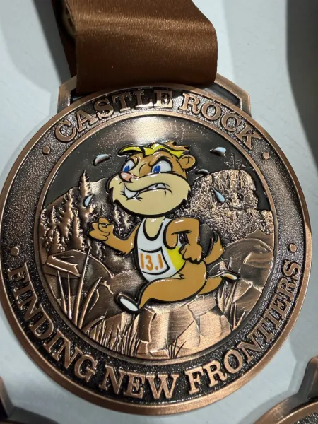 A close-up photo of a bronze medal for the Castle Rock Finding New Frontiers race, featuring a cartoon prairie dog running in a mountainous landscape.