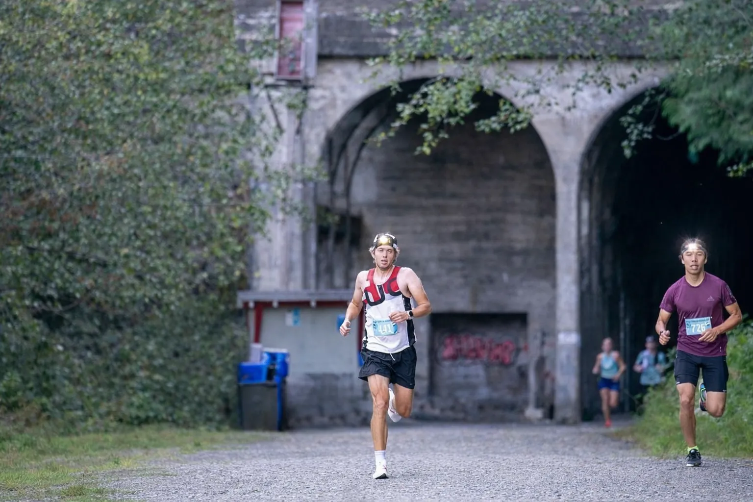 Runners on a gravel path with arched concrete structures in the background.