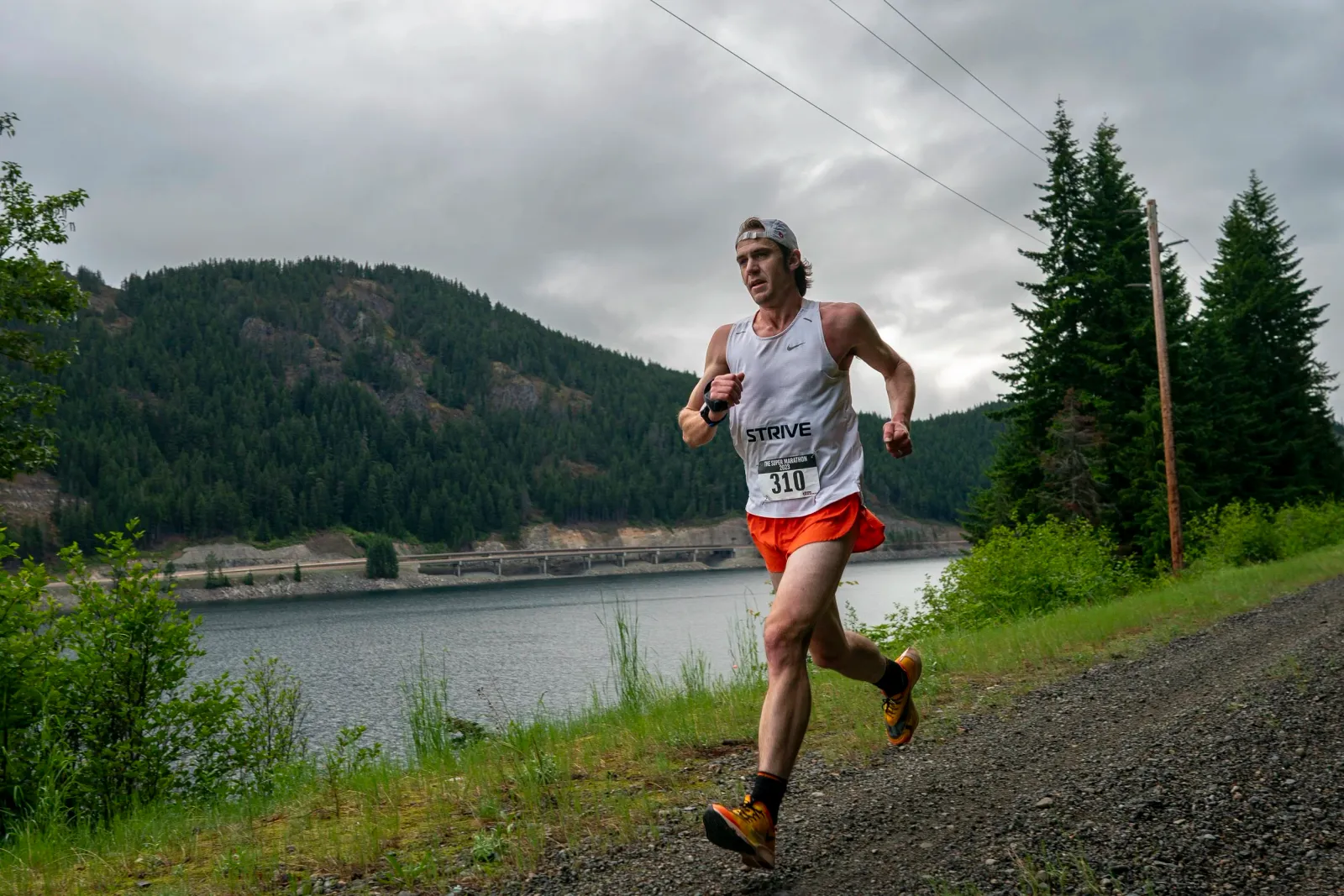 A male runner in a white tank top and orange shorts runs on a gravel path next to a large body of water with forested hills in the background.