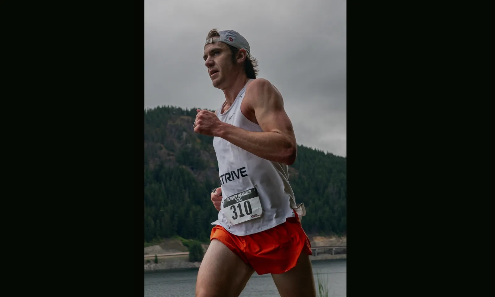A male runner wearing a white tank top with the number 310 and orange shorts runs along a waterfront with a tree-covered hillside in the background.