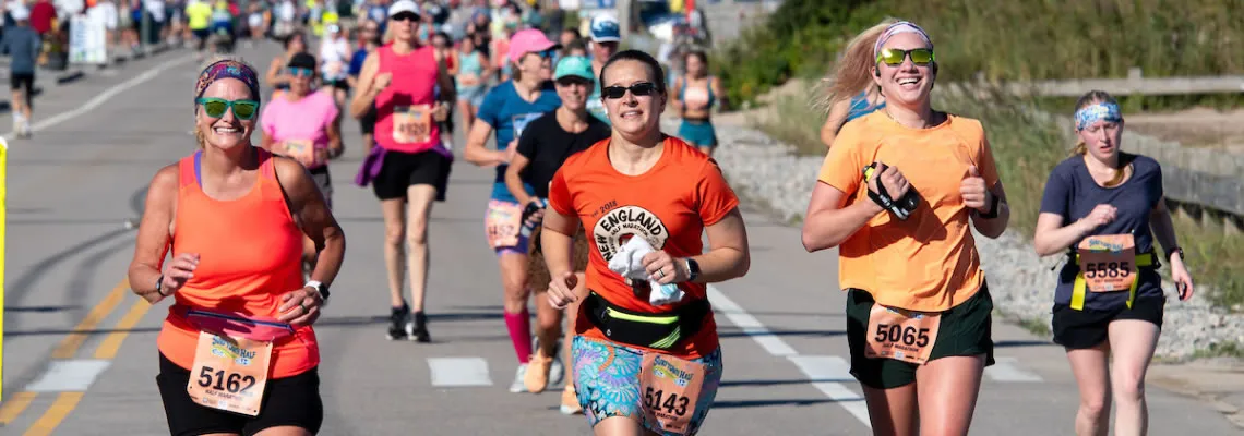 A group of smiling female runners in colorful athletic wear participating in a road race, with the ocean or a beach visible in the background.