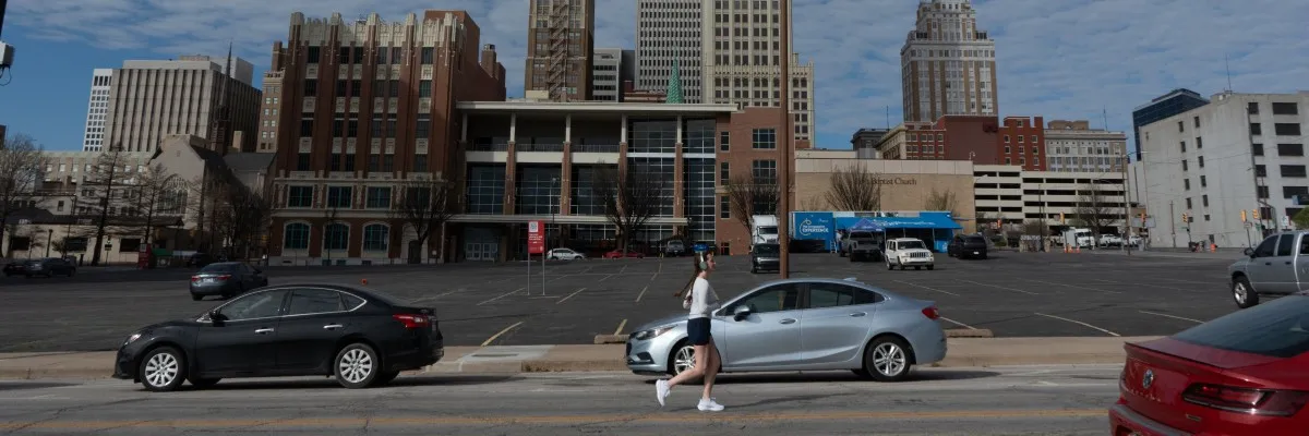 A female runner with headphones runs across a street in an urban environment with tall buildings in the background.