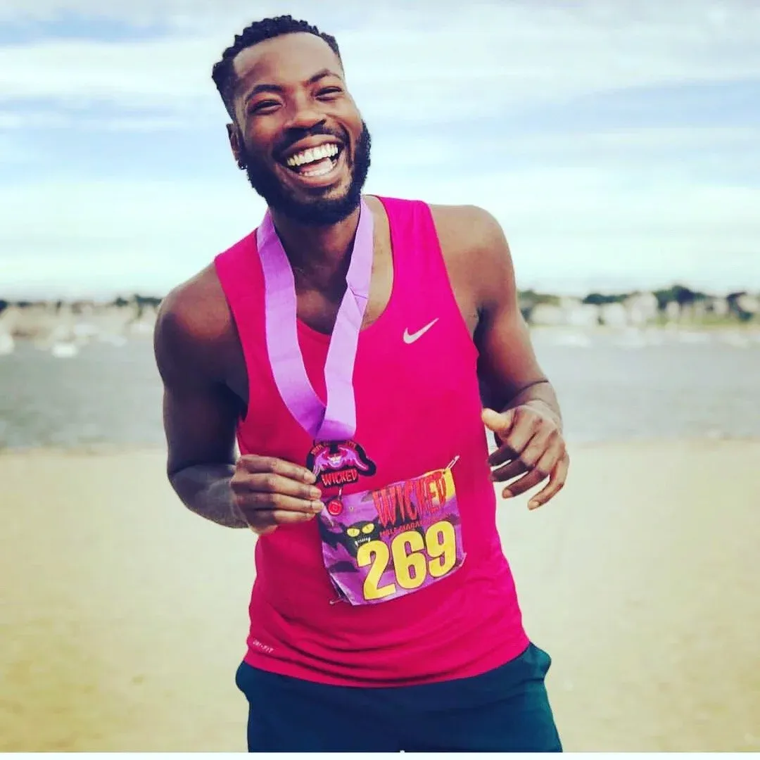 A man wearing a pink tank top and a medal smiles broadly, holding up his race bib with the number 269.