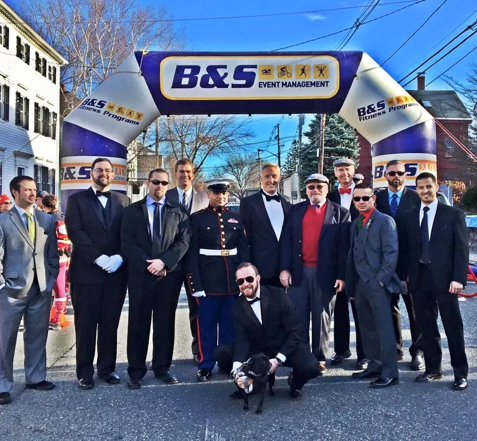 A group of men in formal attire and a military uniform pose under a B&S Event Management archway at a race event.