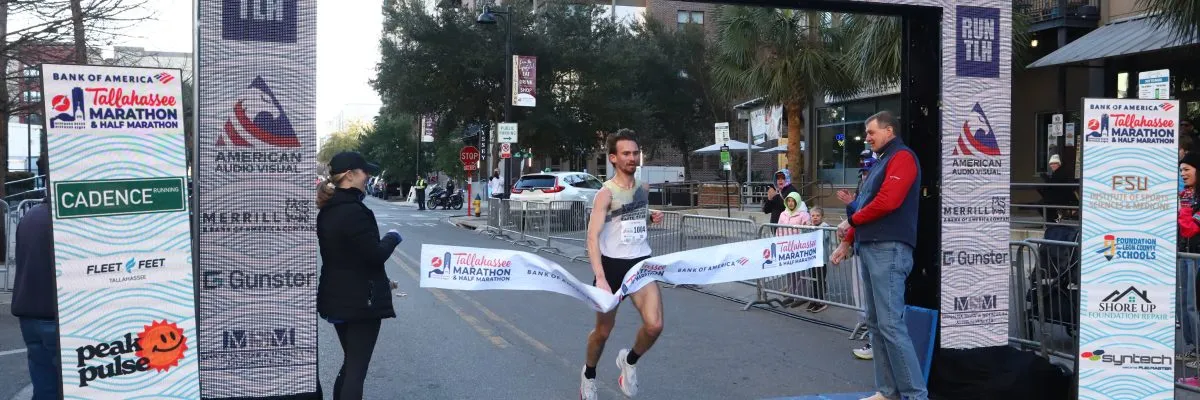A male runner crosses the finish line tape at the Tallahassee Marathon & Half Marathon, with spectators and race officials visible.