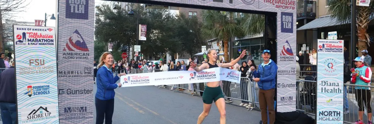 A female runner joyfully breaks through the finish line tape at the Tallahassee Marathon & Half Marathon, with spectators and race officials visible in the background.