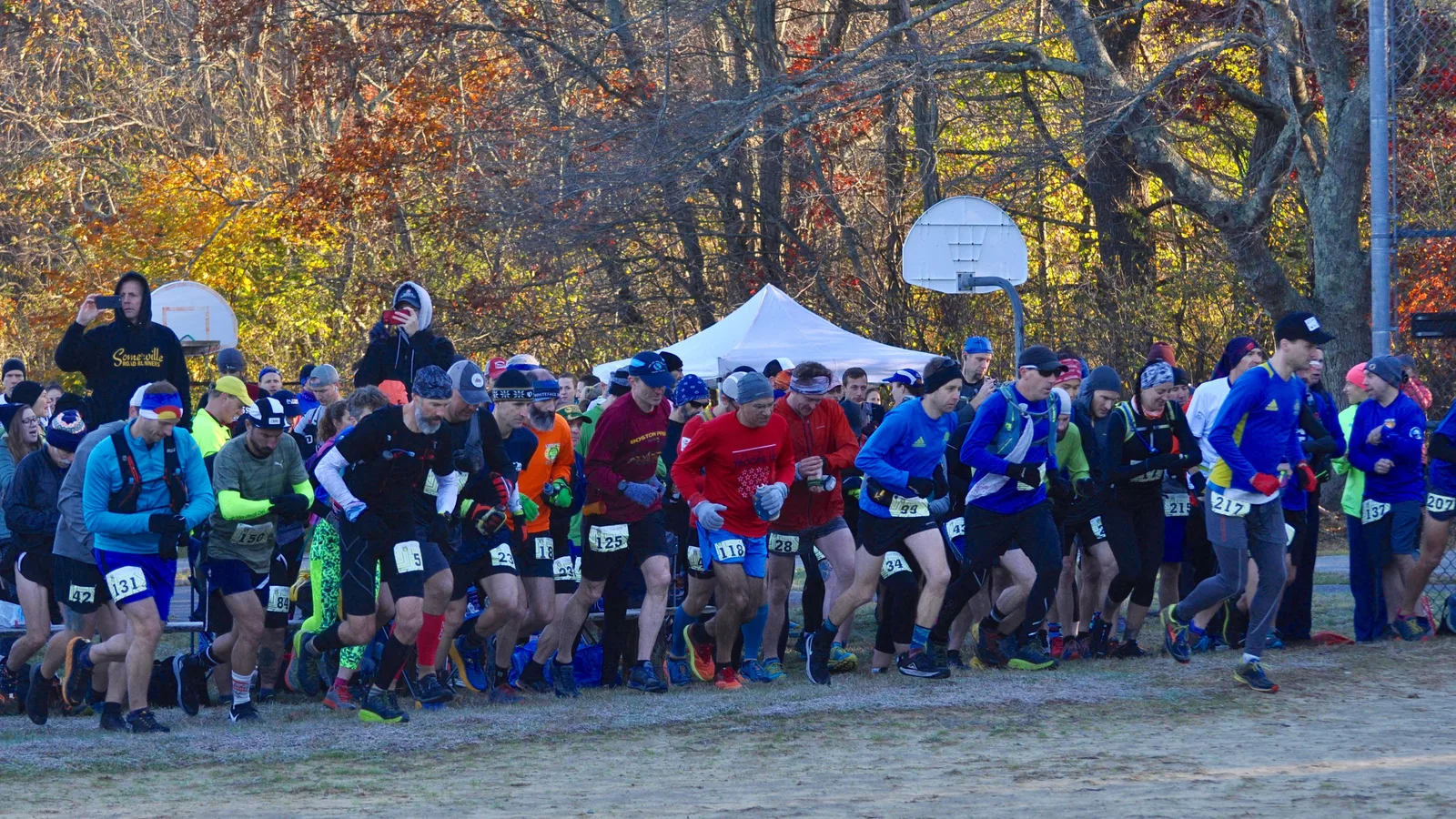 A group of runners in colorful racing attire are starting a race, with trees and a basketball hoop in the background.