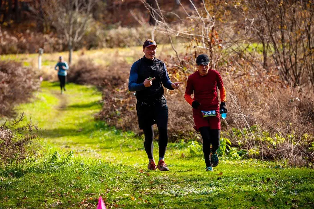 Two runners are on a grassy path with trees in the background, one pointing and talking to the other.