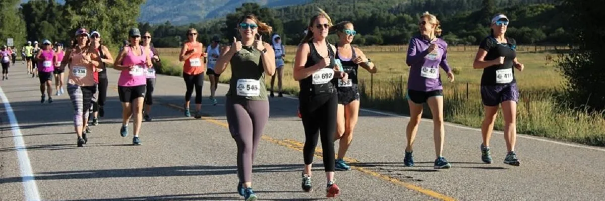 A group of women running a half marathon on a paved road with scenic mountains and trees in the background.