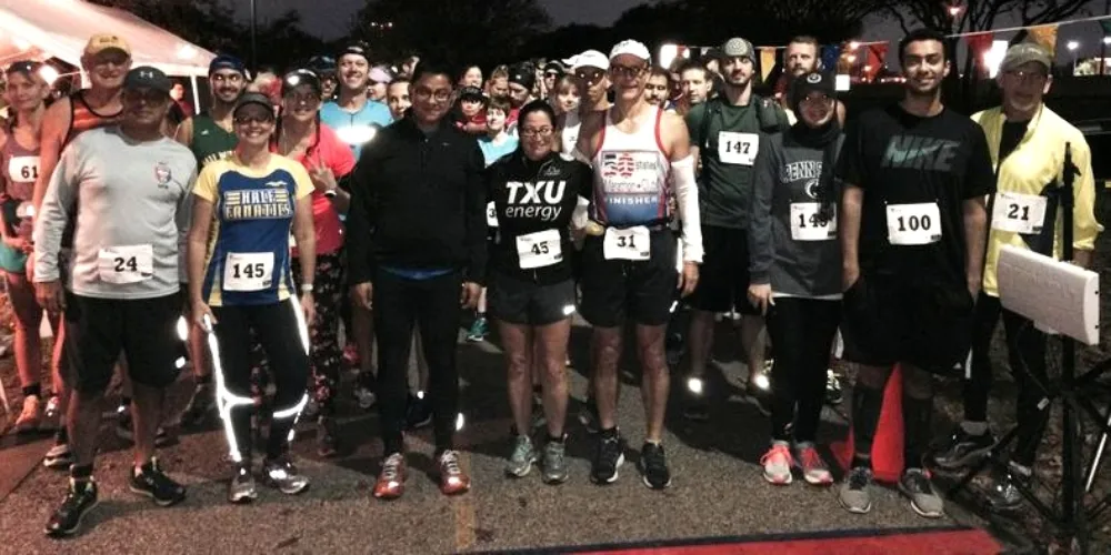 A group of runners stand together at the start line of a race, some wearing bib numbers.