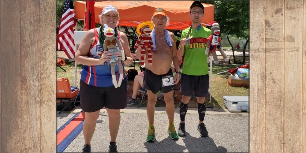 Three runners pose for a photo, two holding sock monkeys and one displaying a medal, with a finish line in the foreground.