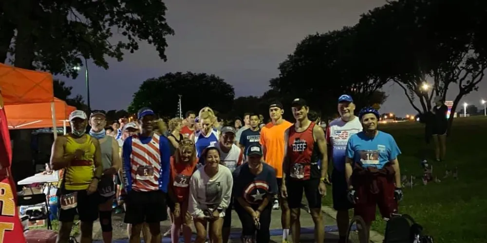 A group of runners stand at the start line of a race at dusk, some wearing patriotic-themed attire.