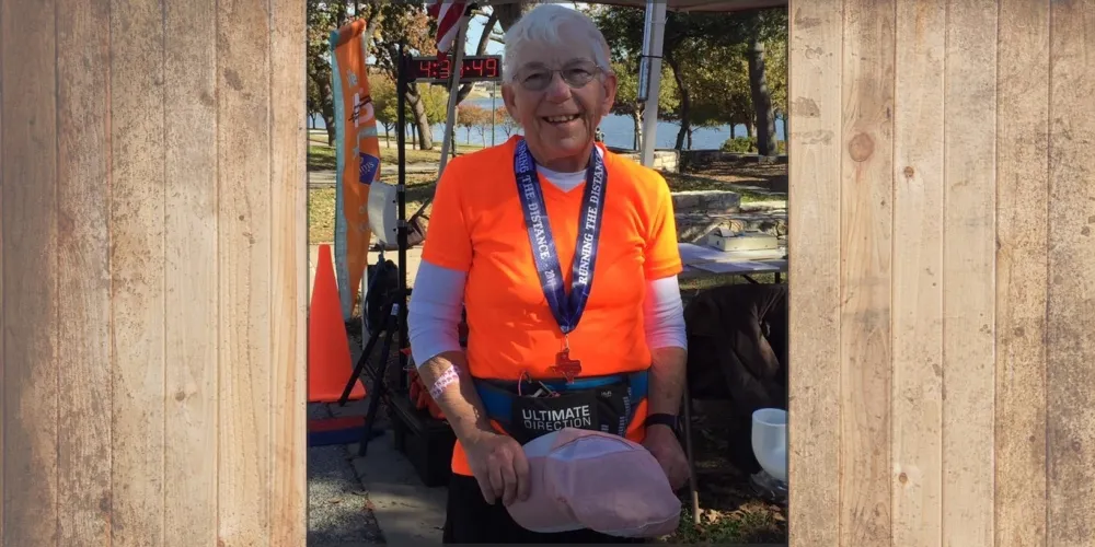 An older woman wearing an orange shirt and a race medal smiles at the camera, holding a hat, with a finish line clock in the background.