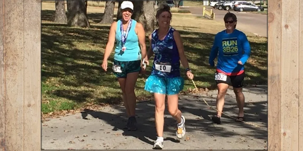 Three female runners smiling as they run on a paved path, with trees and cars in the background.